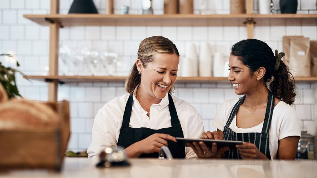 Two women in a kitchen setting laughing, holding an tablet