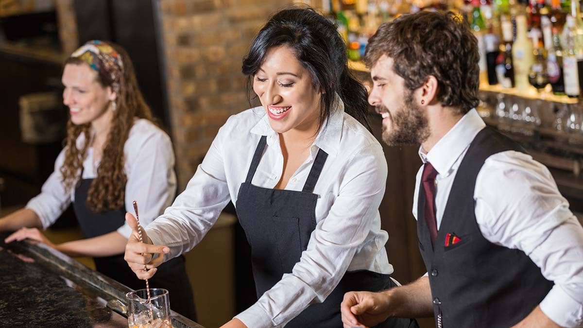 Three bar staff.  One attending to a customer (a woman) at the bar and the others (a man and a woman) making a drink at the bar