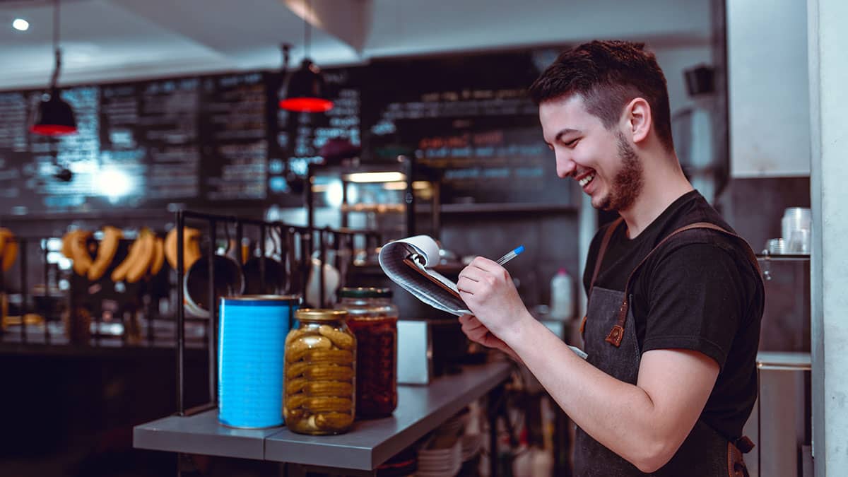 Male hospitality worker behind the counter with a clipboard writing notes 