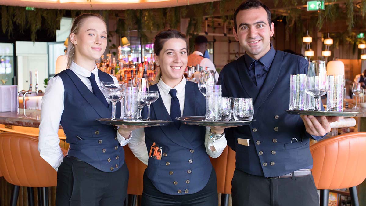 Three happy hospitality staff in uniform, each holding a tray of glasses in front of a restaurant bar.