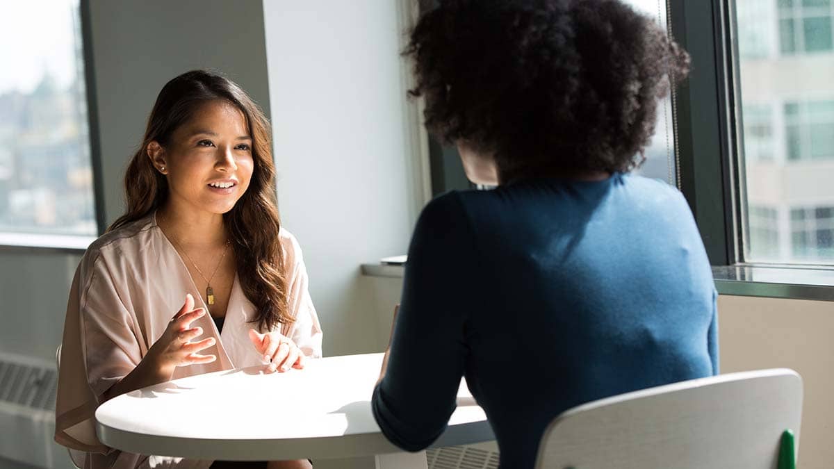 Two women at a table having a discussion. One woman is wearing a pink top and is smiling, whilst other you can only see the back of her head but looks engaged in conversation with the woman in front of her.