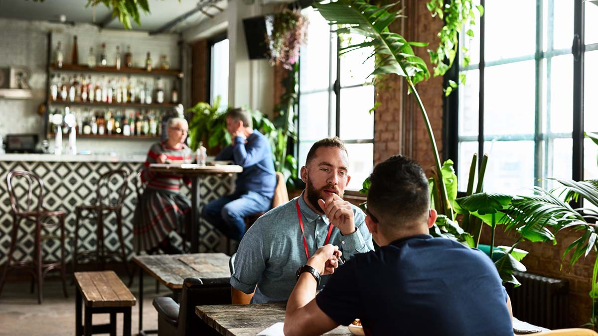 Two men in a restaurant having a conversation.  Blurred in the background is a man and a woman sitting at a separate table also having a conversation.